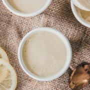 Individual servings of lemon posset sit in white ceramic cups on brown cloth with one copper spoonful resting on one serving.