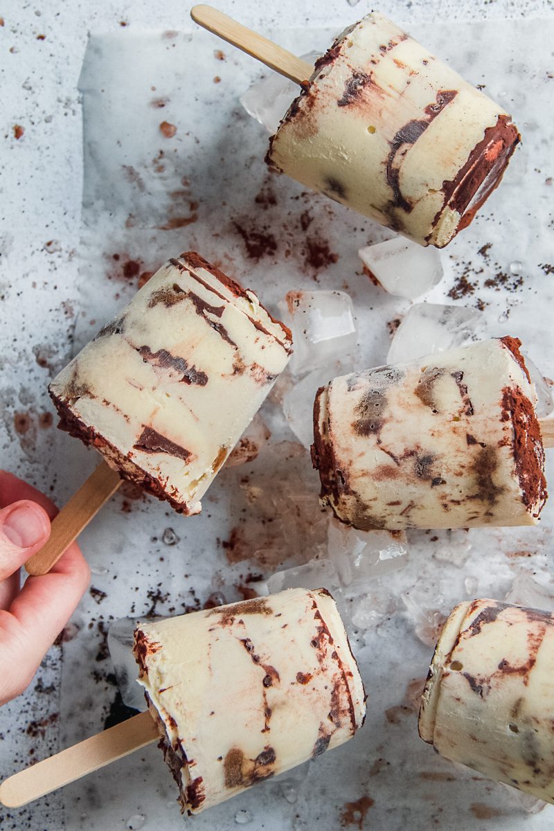 A frozen tiramisu posicle is picked up with other popsicles resting on ice cubes on a light gray surface.