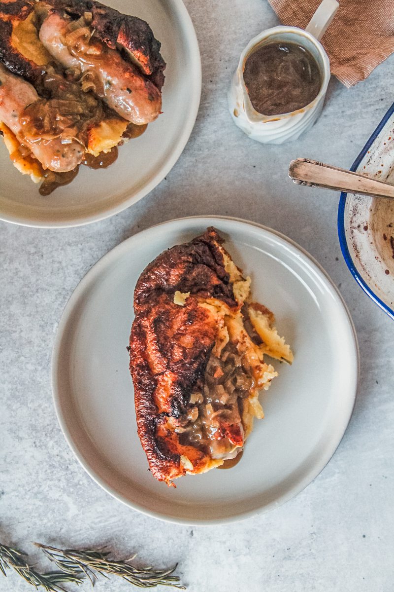 A serving of Toad in a hole sits on rimmed ceramic plates on a gray surface beside a second serving topped with gravy.