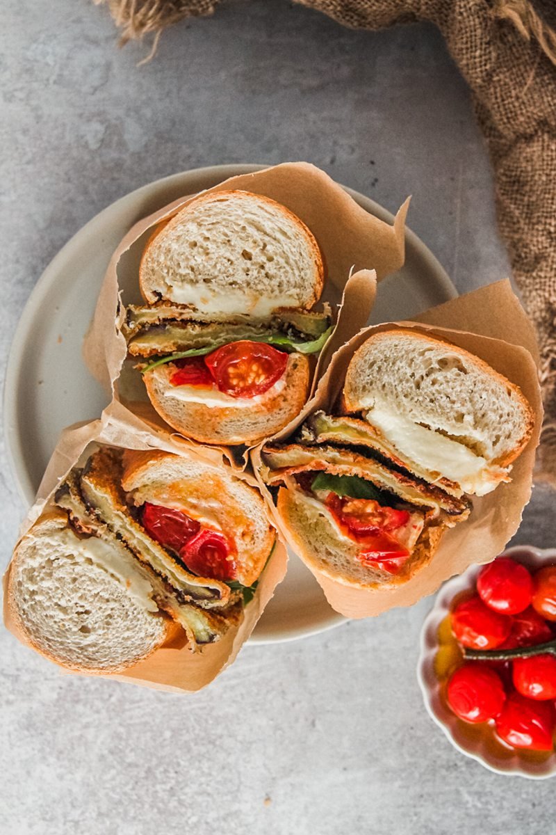 Roasted tomato and eggplant sandwiches sit wrapped in parchment paper on a rimmed ceramic plate on a gray surface.