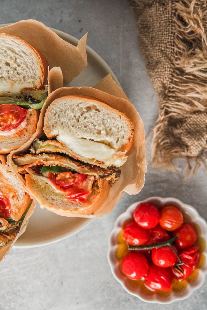 A close up of a roasted tomato, eggplant and mozzarella sandwich sitting on a rimmed ceramic plat on a gray surface with a serving of roasted cherry tomatoes on the side.