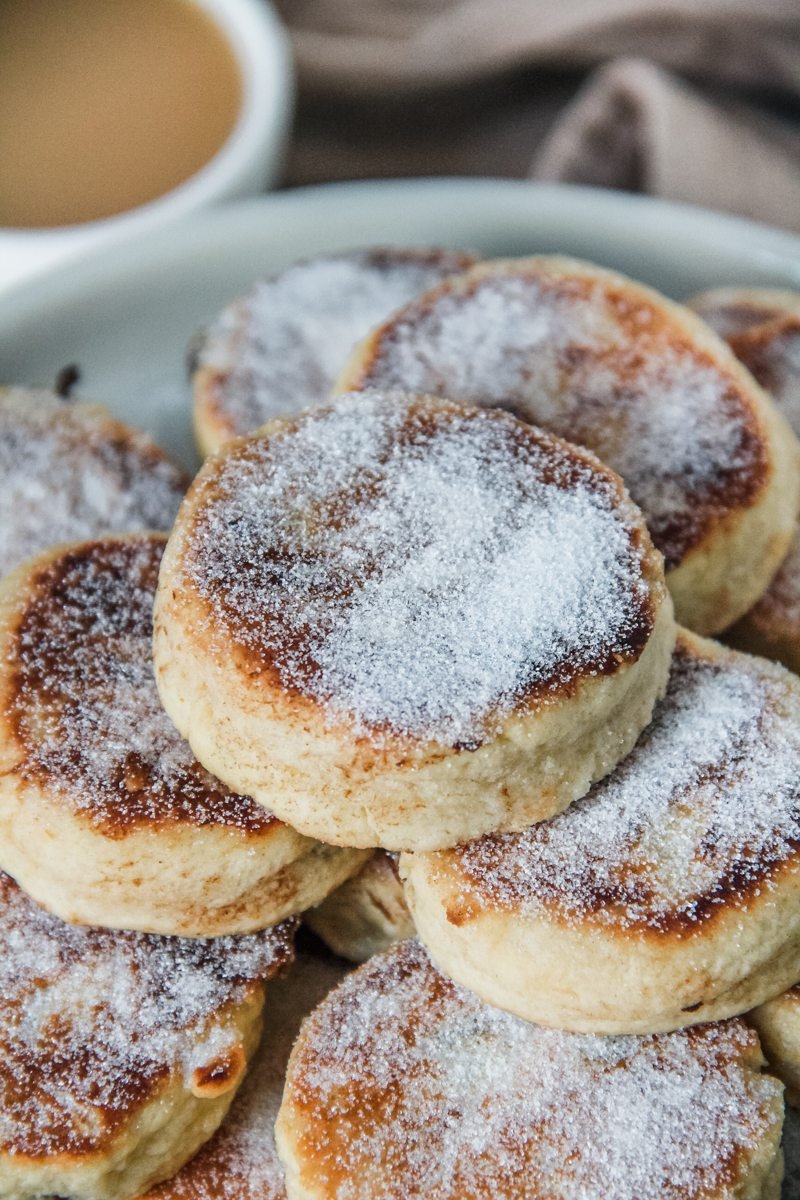 A stack of Welsh cakes sprinkled with sugar sit on a rimmed ceramic plate with a cup of tea visible in the background.