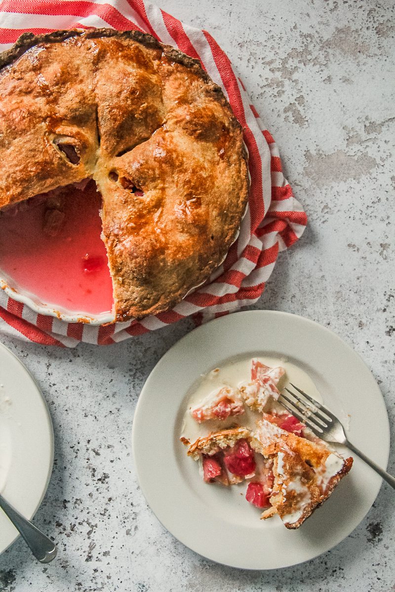 A slice of rhubarb pie with pouring cream poured over and a fork resting on an individual plate sits beside the remaining pie on a striped cloth on a light gray surface.