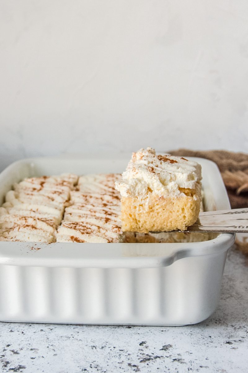 A slice of Tres Leches sits on a cake server on a rectangle white baking ceramic dish on a light gray surface.