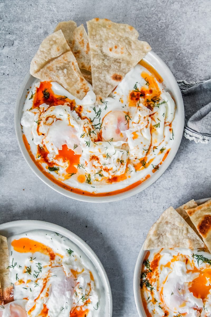 An overhead shot of Cilbir - Poached Eggs in Yogurt on a rimmed ceramic plate with a few pieces of bread served on the side on a gray surface.