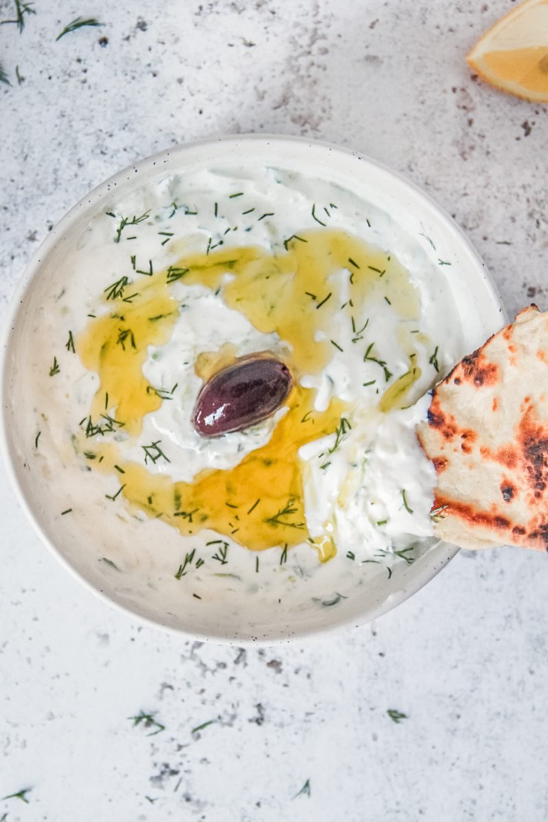 A piece of pita picks up Greek tzatziki from a white ceramic bowl on a light gray surface garnished with a traditional black olive.