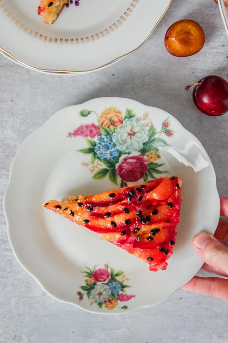 A slice of upside down plum passionfruit cake sits on an individual plate with someone holding the plate above a gray surface.