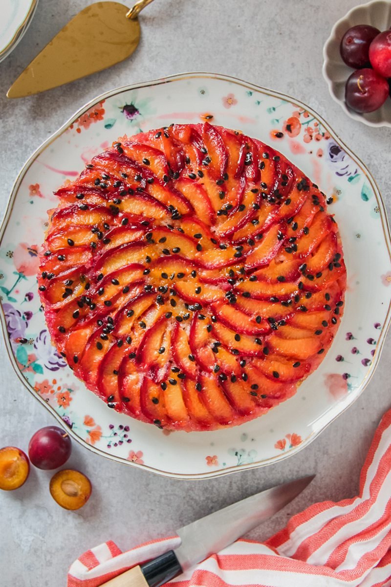 An upside down plum passionfruit cake sits on a floral serving plate on a gray surface.