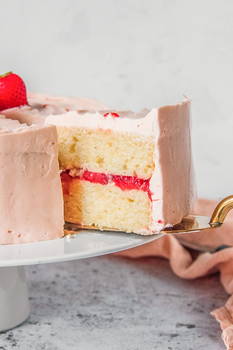 A slice of vanilla strawberry cake sits on a cake server on a light gray surface.