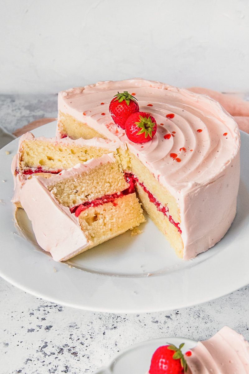 Slices of vanilla strawberry cake with a roasted strawberry center and strawberry Swiss meringue buttercream sits on a white ceramic plate on a light gray surface.