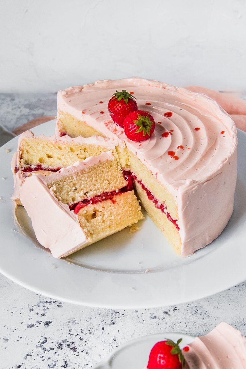 Slices of vanilla layer cake with a roasted strawberry center and strawberry swiss meringue buttercream frosting sits served up on a white plate on a light grey background with two slices leaning against each other on the serving plate.