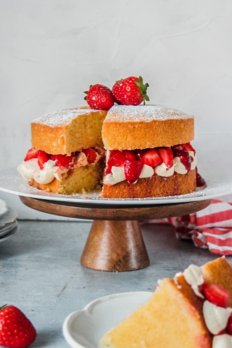 A sliced Victoria sponge cake sits on a white ceramic plate on a wooden cake stand on a gray surface.