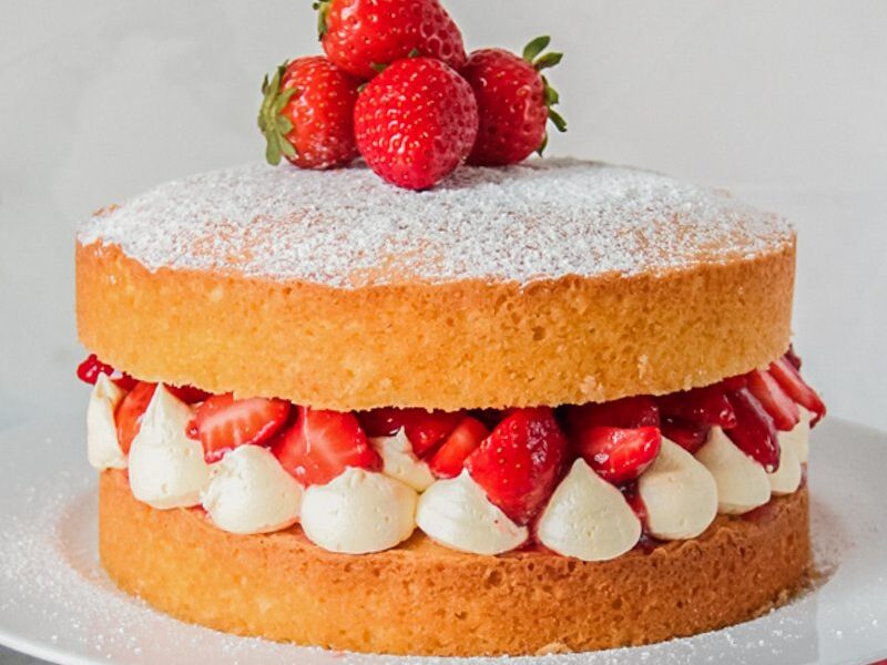 Victoria sponge cake sits on a white ceramic plate on a wooden cake stand on a gray surface.
