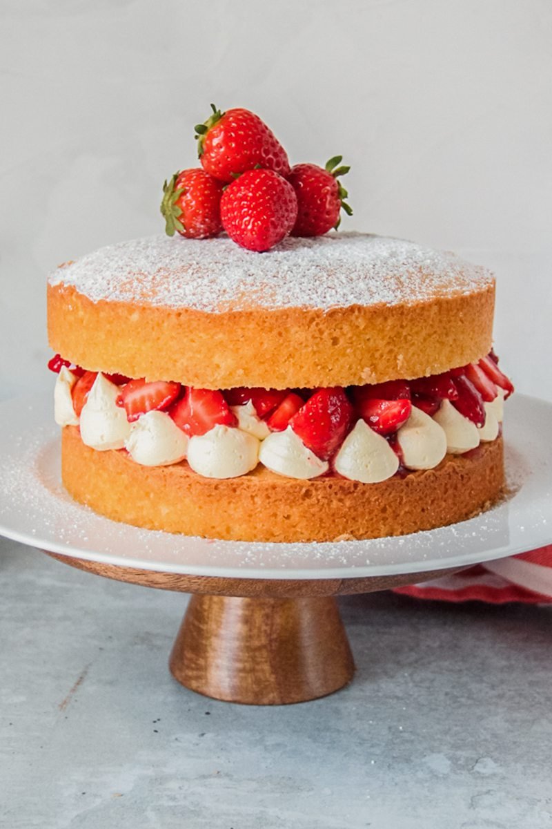 Victoria sponge cake sits on a white ceramic plate on a wooden cake stand on a gray surface.