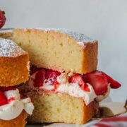 A slice of Victoria sponge cake sits a cake server on a white ceramic plate on a wooden cake stand on a gray surface.