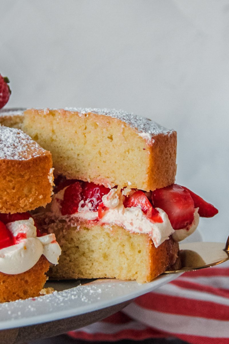 A slice of Victoria sponge cake sits a cake server on a white ceramic plate on a wooden cake stand on a gray surface.