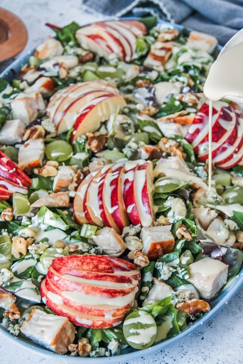 A drizzle of dressing is poured over waldorf salad on a blue plate on a gray surface.