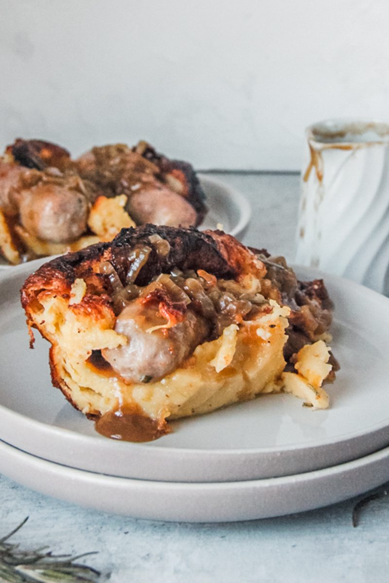 An individual serving of Toad in the hole sits on a stack of rimmed ceramic plates topped with a caramelized onion gravy on a gray surface.