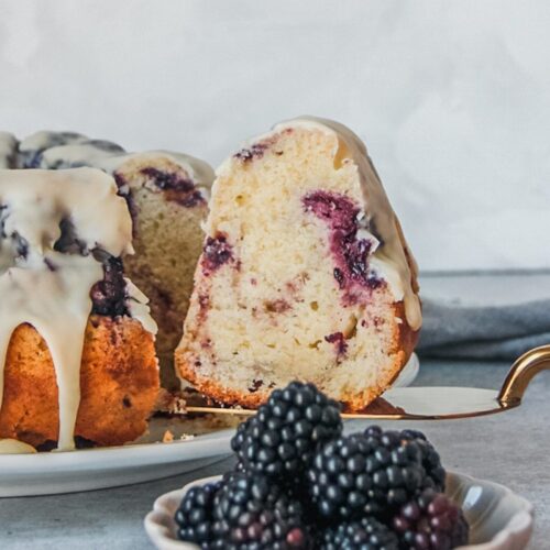 A slice of white chocolate blackberry cake sits on a cake server beside the remaining cake with blackberries on a small plate in front of the slice on a gray surface.