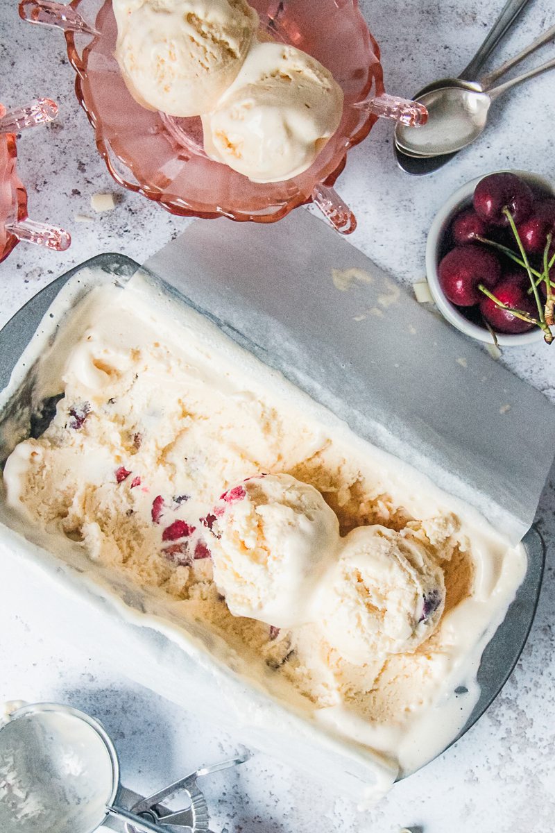 Scoops of white chocolate cherry semifreddo sit on top of a lined rectangle metal baking tin with pink glass bowls on a light gray surface.