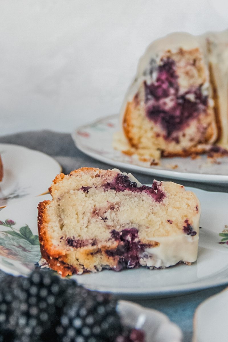 A slice of white chocolate blackberry bundt cake sits on an individual plate on a gray surface with the remaining cake in the background.
