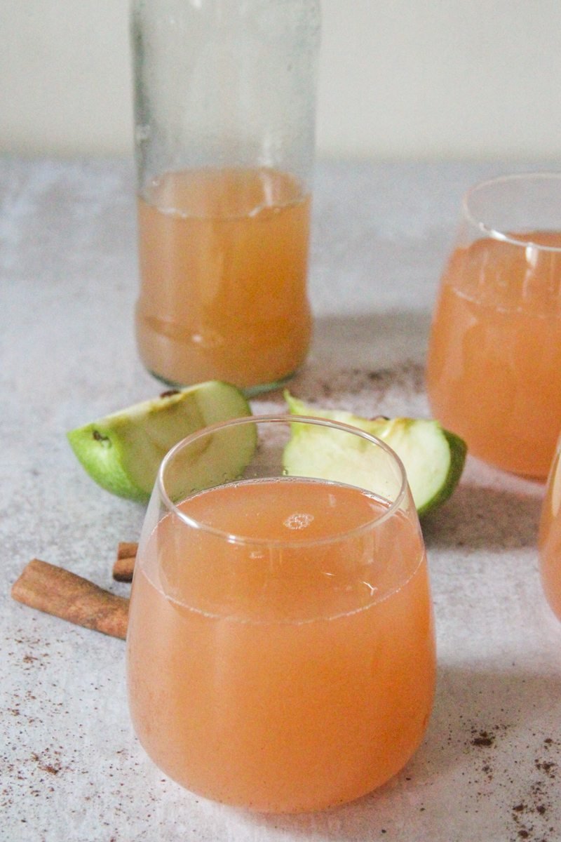 Low ball glasses sit on a gray surface with golden homemade apple cider inside and quartered apple and cinnamon sticks in the background.