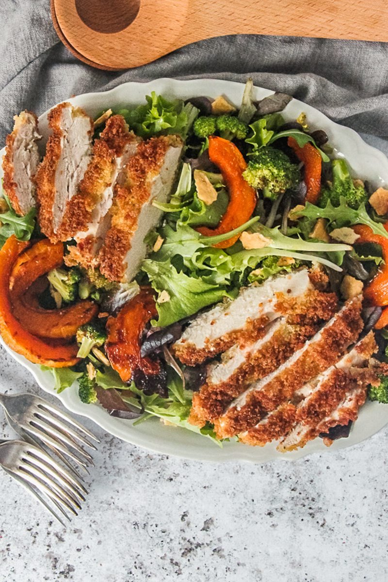 Breaded crispy chicken slices on top of lettuce and roasted butternut squash served up on a white plate on a gray surface with forks and salad servers on the side.